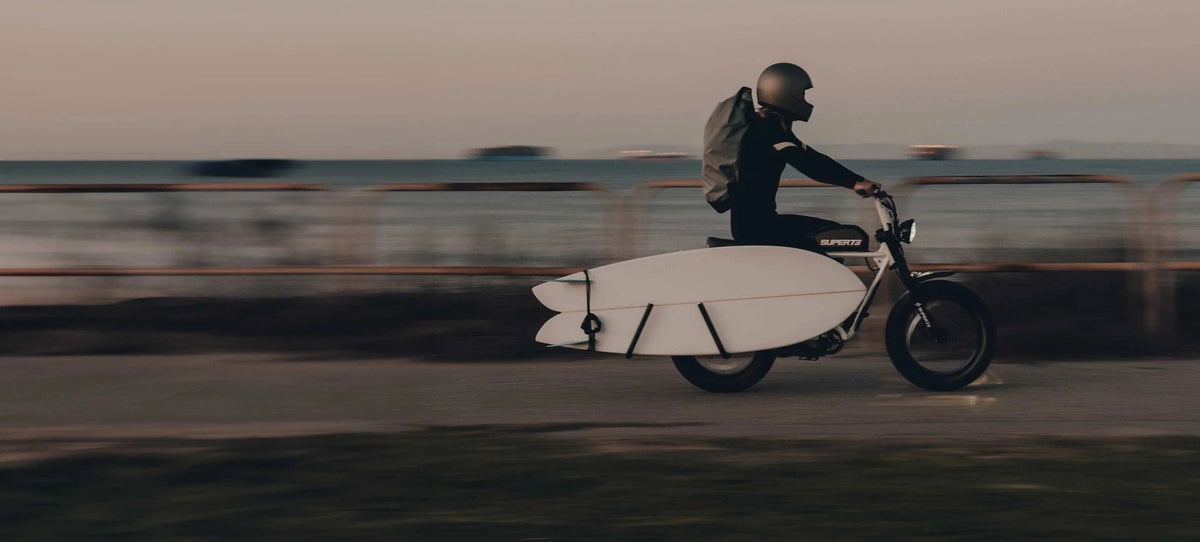 Surfer in a helmet riding a Super73 e-bike with a surfboard on a side rack along a beachfront path at dusk