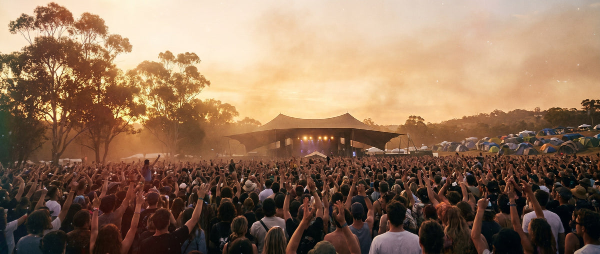 Festival crowd with arms raised in front of an outdoor stage at sunset, gum trees to one side and a campsite of tents on the hill behind